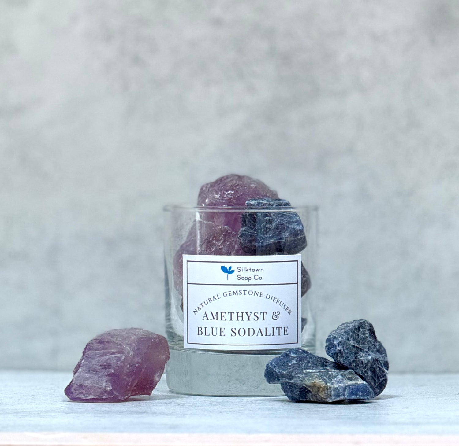 A natural gemstone diffuser jar on a white surface containing raw, unpolished Amethyst and Blue Sodalite gemstones with a label indicating the types of gemstones. Additional Amethyst and Blue Sodalite gemstones are placed outside of the jar.