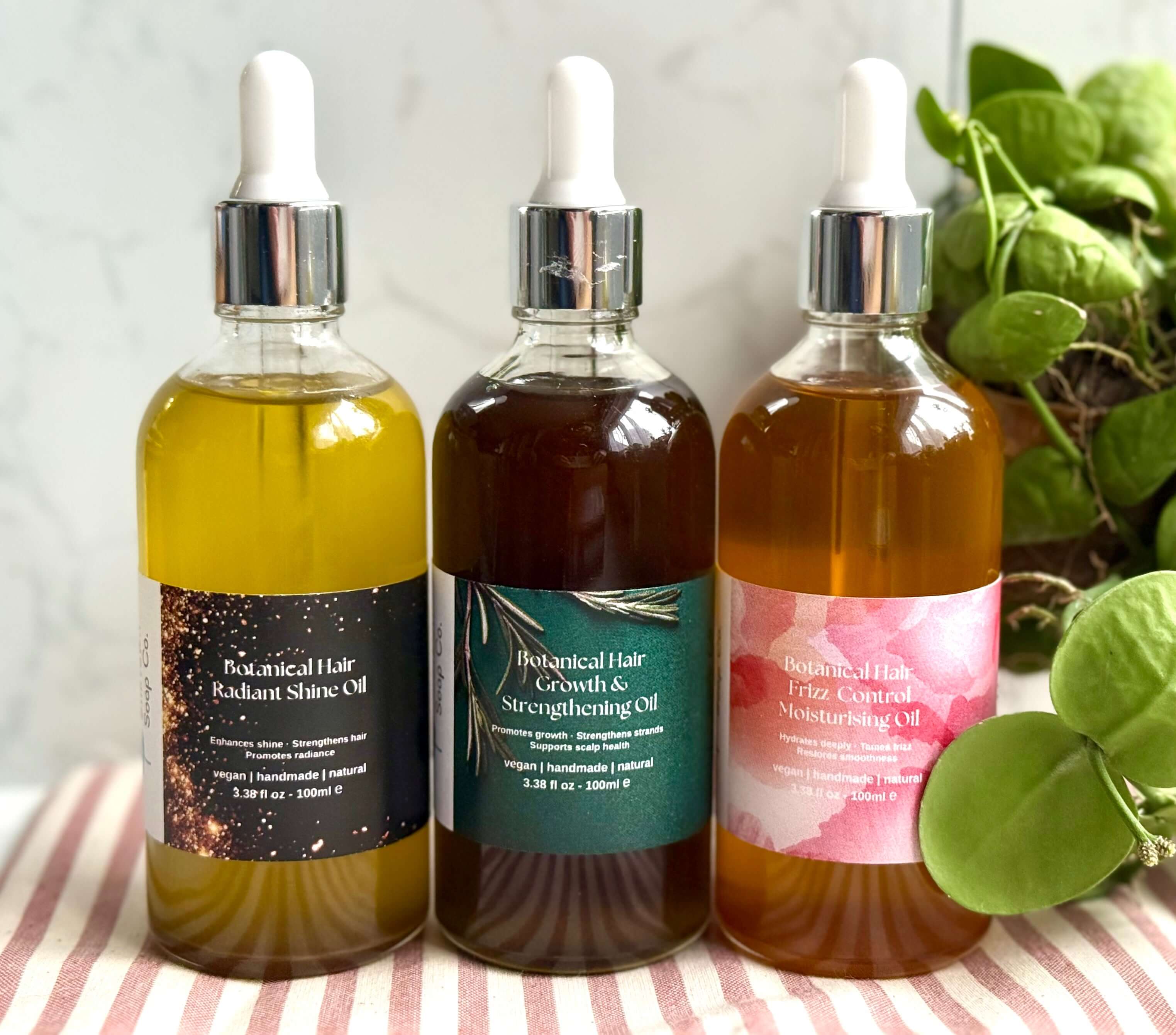 Three bottles of botanical hair care oils with labels on a striped fabric background.