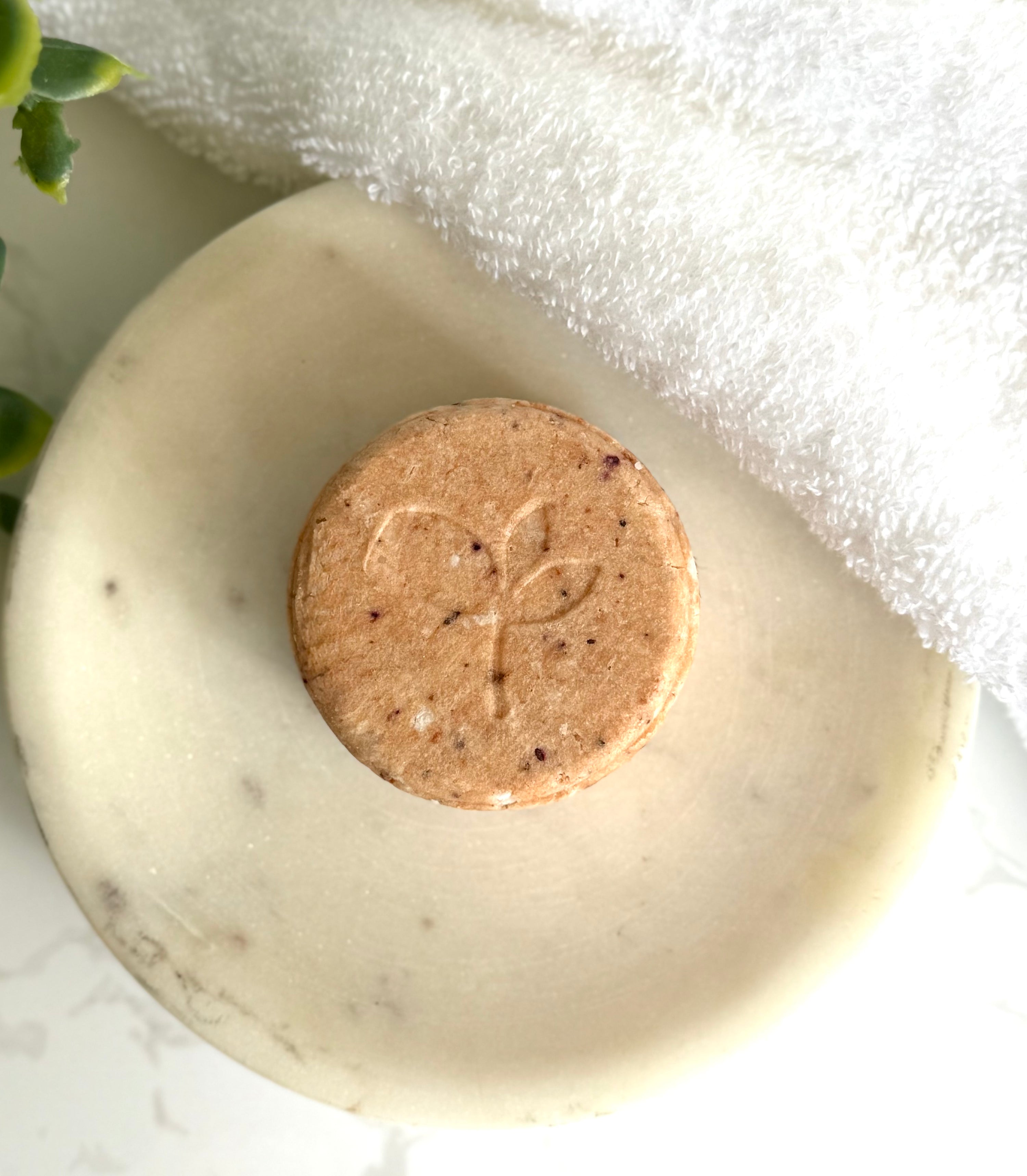 Bar of Natural Shampoo Bar - Good Hair Day with a circular stamp on a white dish with a towel and foliage in the background