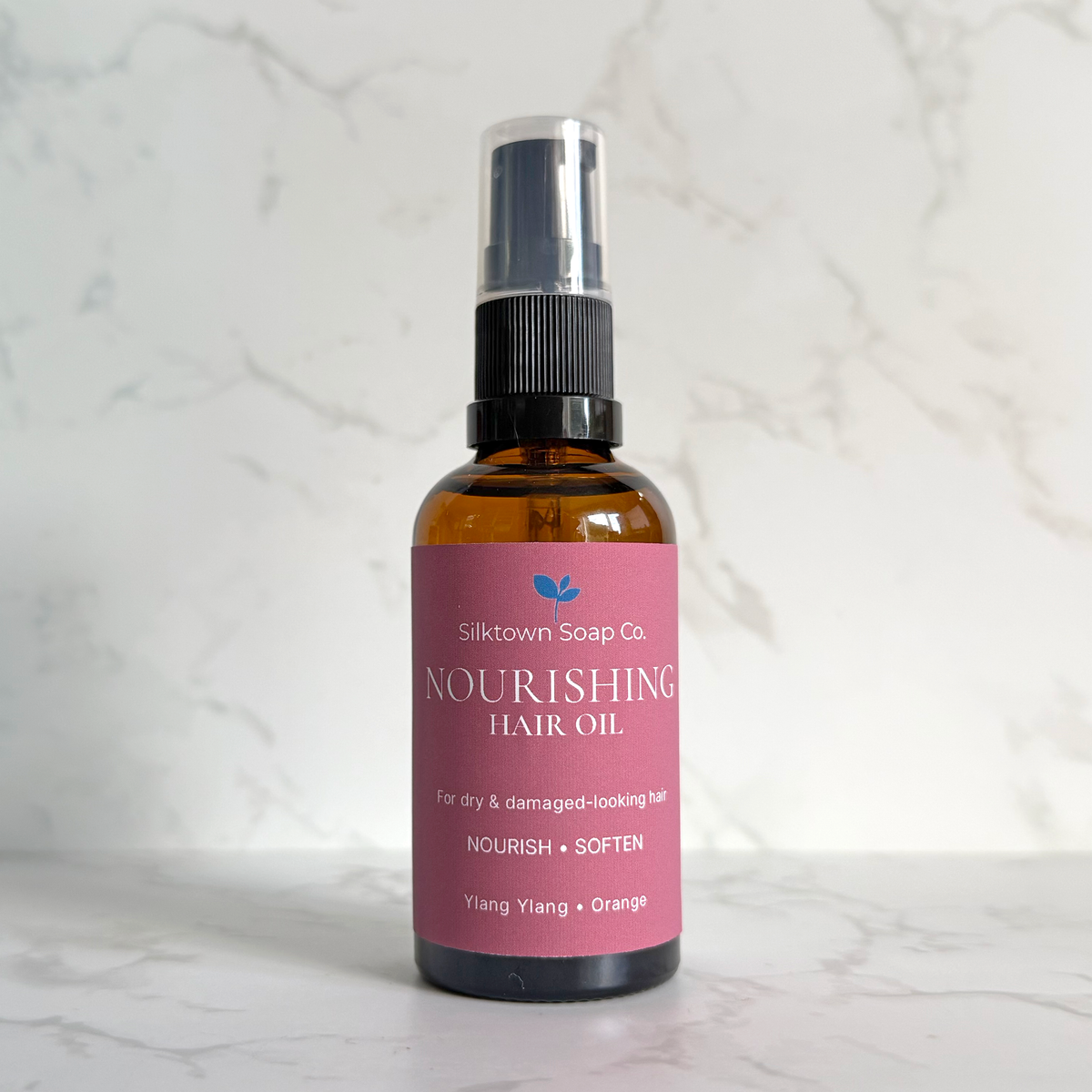 A bottle of Silktown Soap Co. Nourishing Hair Oil in the Ylang + Orange scent, featuring an amber glass bottle with a pink label and a black spray nozzle, set against a white marble background.