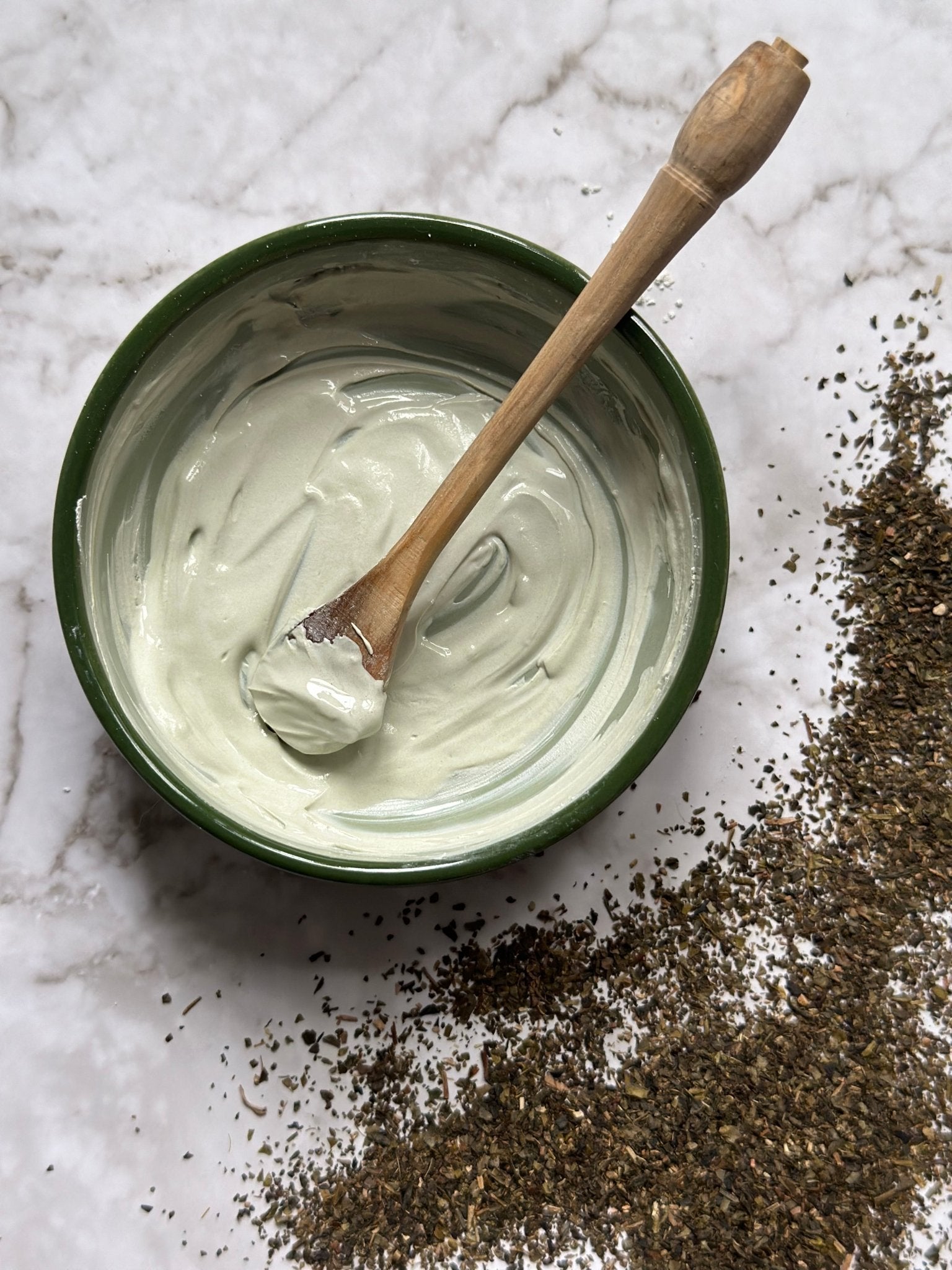 Hydrate Natural Clay Mask in a bowl with a wooden spoon after being mixed on a marble background with green tea granules