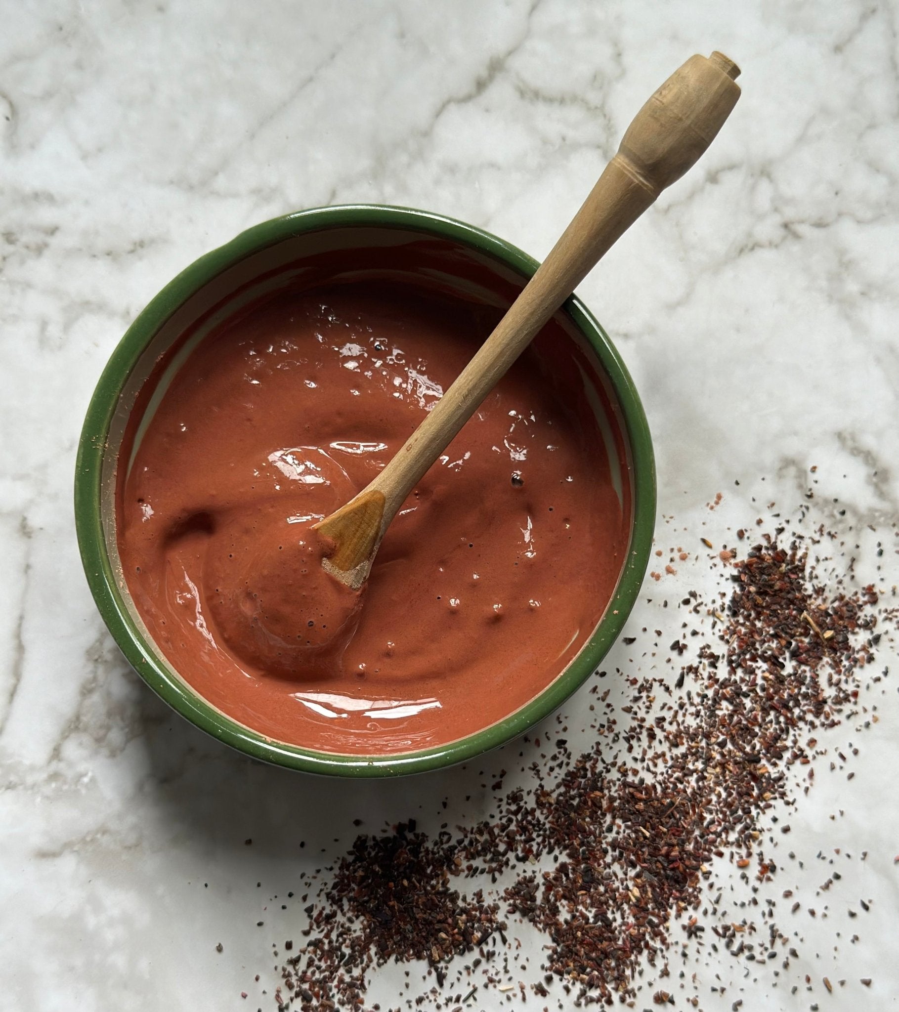 Recharge Natural Clay Mask in a bowl with a wooden spoon after being mixed on a marble background with hibiscus granules