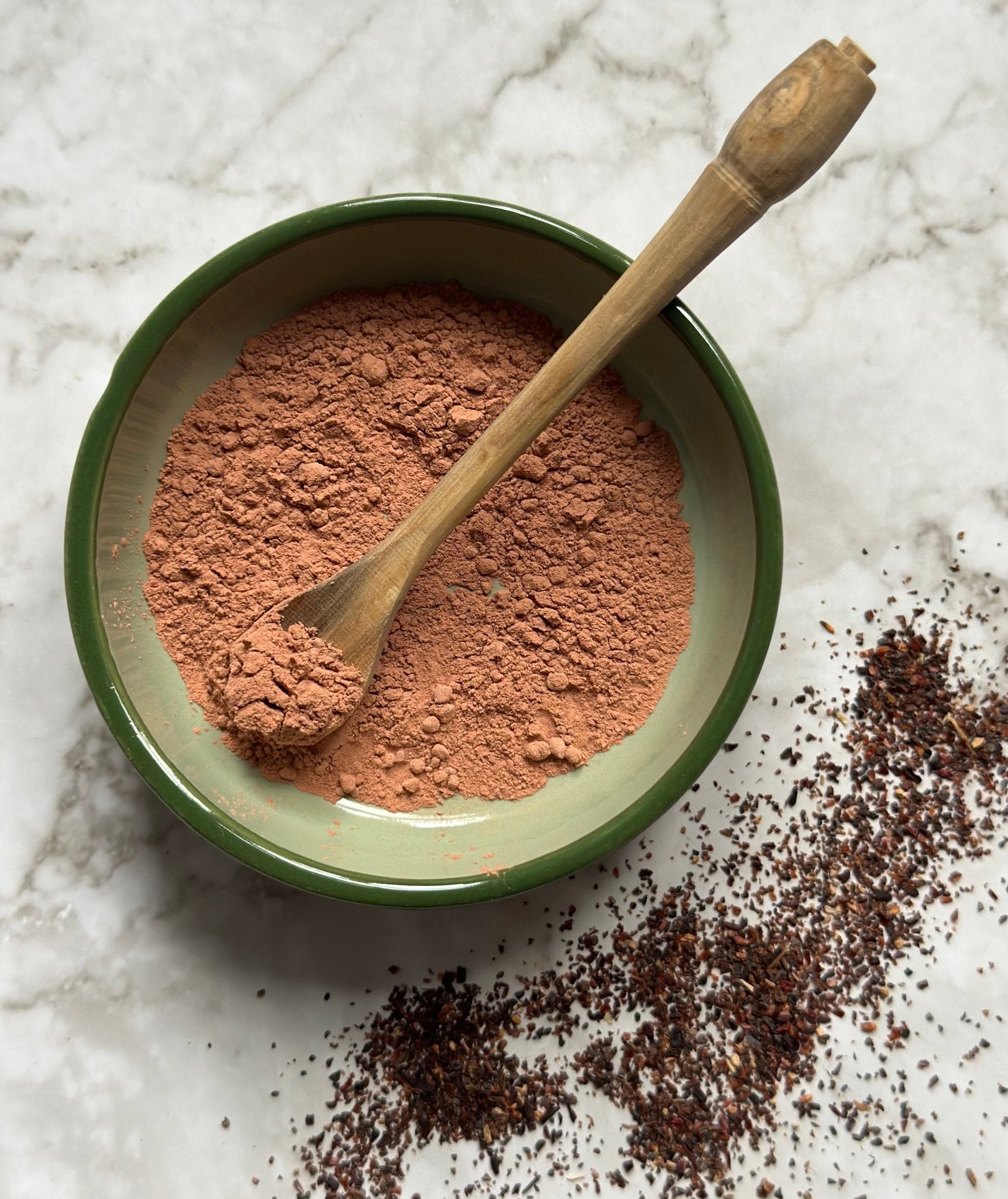 Recharge Natural Clay Mask in a bowl with a wooden spoon before being mixed on a marble background with hibiscus granules