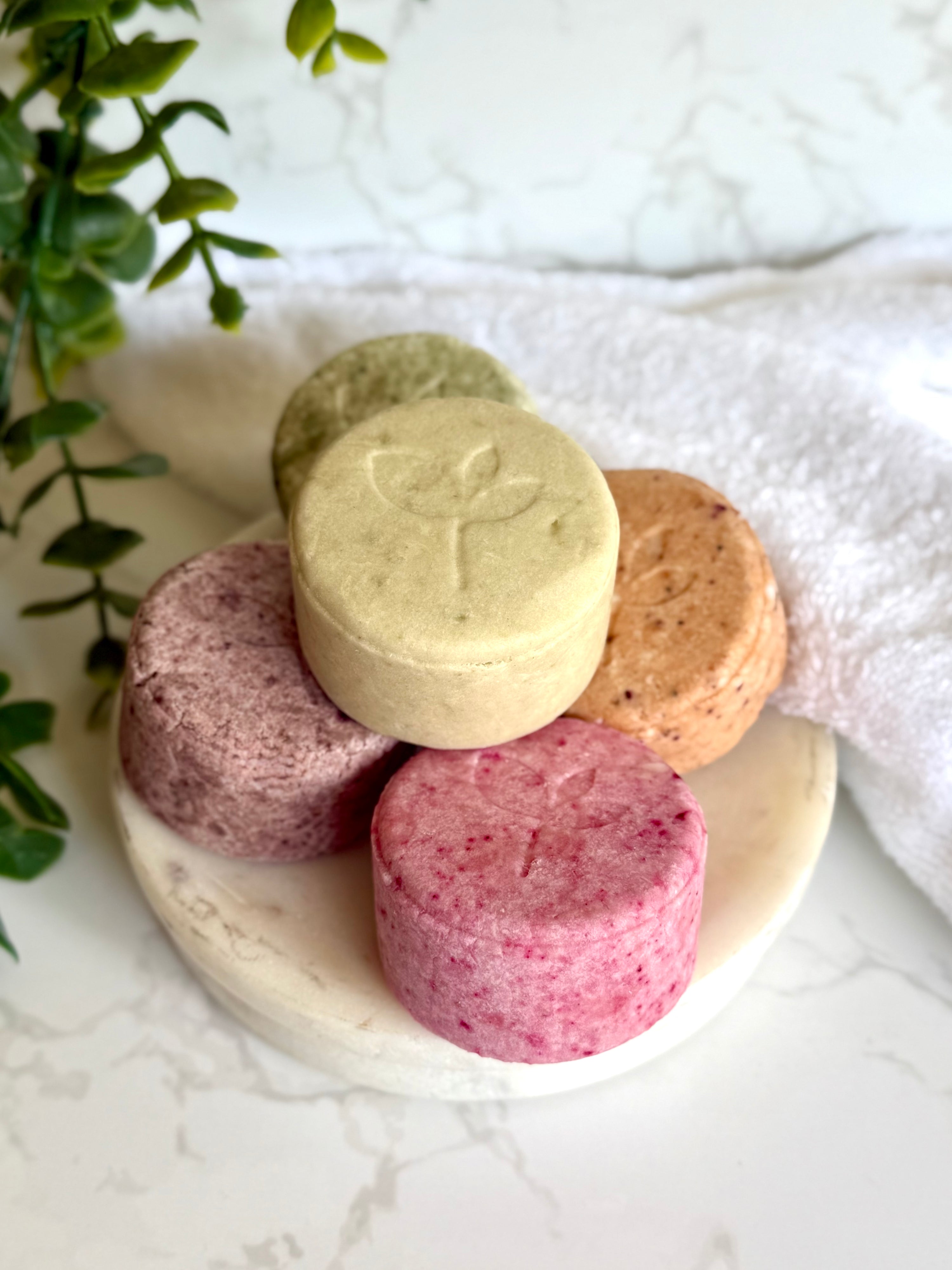 Five colorful natural shampoo bar on a marble surface with a white towel and green leaves in the background.