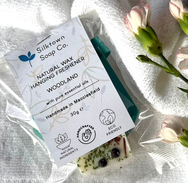 A packaged natural wax freshener with woodlands aroma, adorned with dried botanicals, and labeled as handmade in Massachusetts, 100% natural vegan wax, and eco-friendly.