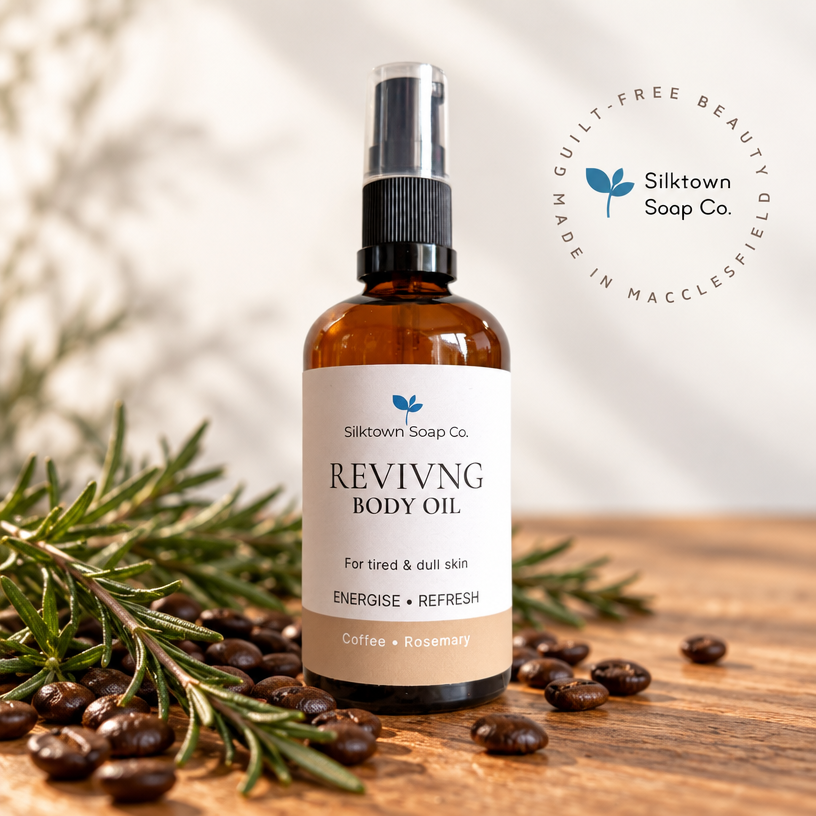 Amber glass spray bottle of Silktown Soap Co. Revivng Body Oil with a white label, styled on a wooden surface with coffee beans and rosemary sprigs, with a circular badge in the top right.