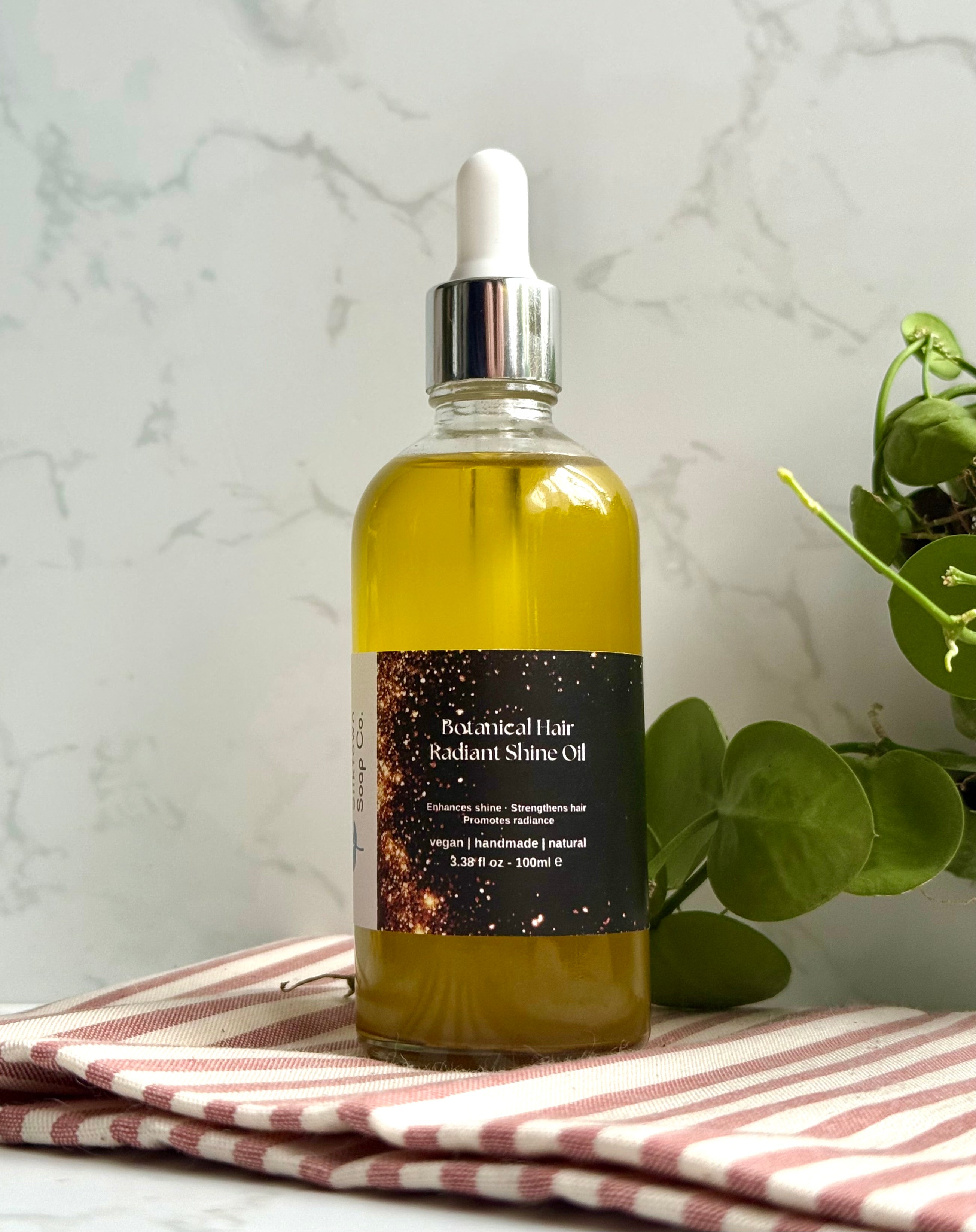 Bottle of Botanical Hair Radiant Shine Oil on a striped cloth with a plant in the background