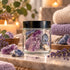 A clear glass jar with a black lid, filled with purple and blue crystals, set on a marble surface with lavender, candles, and folded towels in the background.