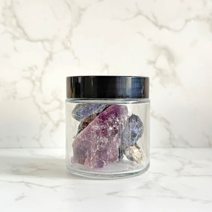 A clear glass jar with a black lid containing several rough crystals, including a prominent purple crystal, set on a white marble surface against a matching marble background.