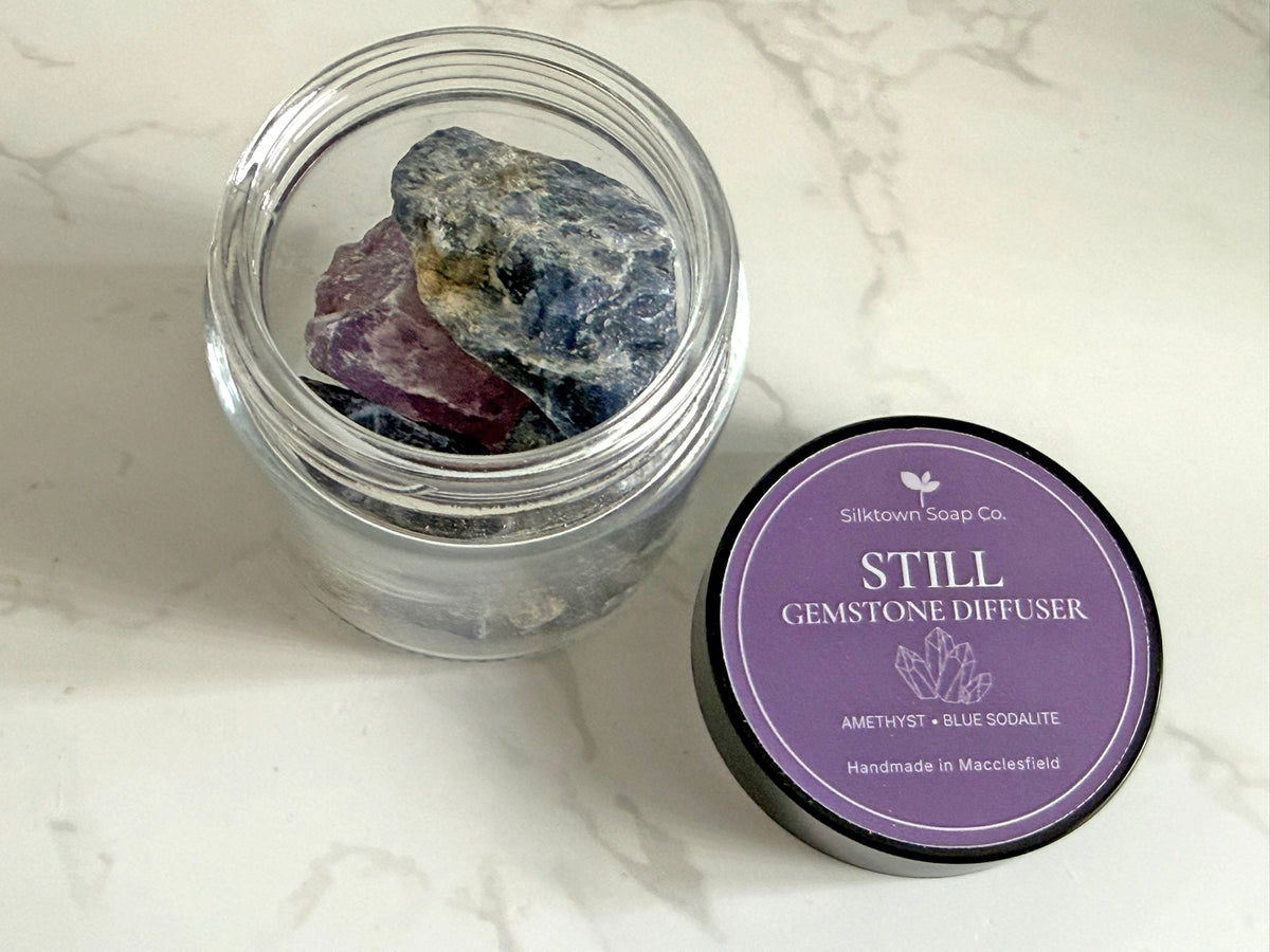 An open clear glass jar filled with gemstones sits on a white marble surface next to its purple lid, which reads &