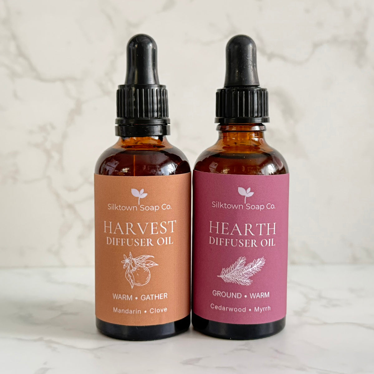Two amber glass dropper bottles of Silktown Soap Co. diffuser oil, labeled Harvest Diffuser Oil and Hearth Diffuser Oil, standing side by side on a light marble surface against a marble background.