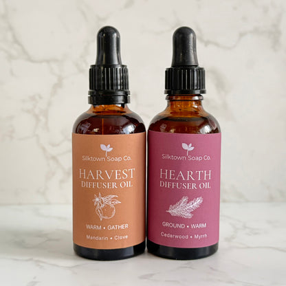 Two amber glass dropper bottles of Silktown Soap Co. diffuser oil, labeled Harvest Diffuser Oil and Hearth Diffuser Oil, standing side by side on a light marble surface against a marble background.