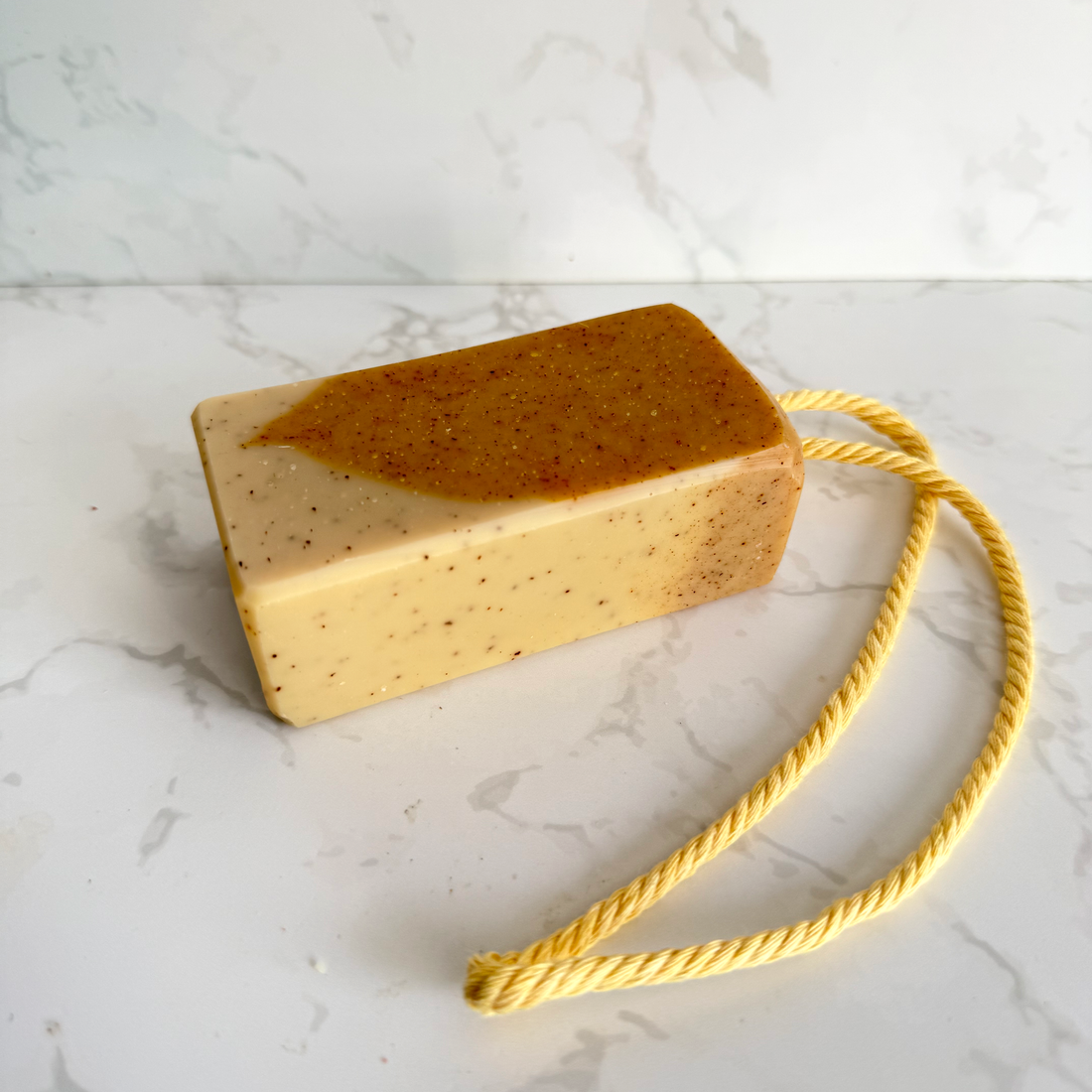 A rectangular bar of handmade soap in a warm beige and orange-brown speckled pattern, featuring a yellow rope tag, set against a white marble background.