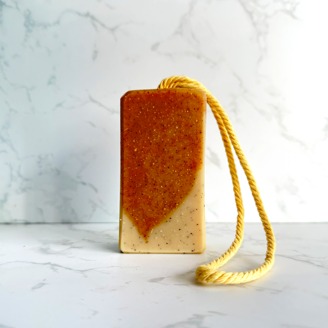 A rectangular handmade soap bar with a yellow rope loop, featuring a two-tone design with a textured orange-brown top half and a smooth beige bottom half, set against a white marble background.