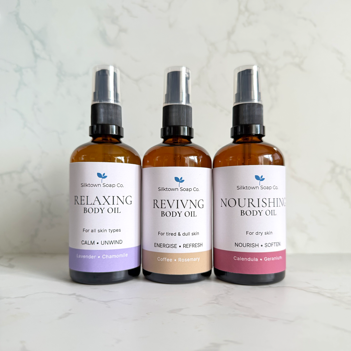 Three amber glass body oil spray bottles from Silktown Soap Co. lined up on a light marble surface against a marble wall.