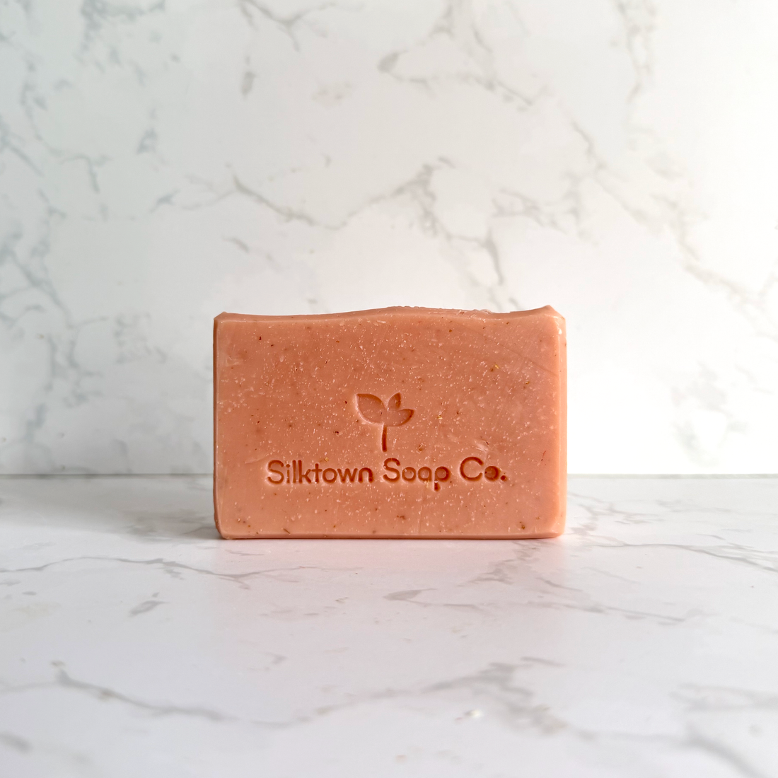 A rectangular bar of Silktown Soap Co. soap in a warm peachy-pink color, standing upright on a white marble surface with a matching marble background.