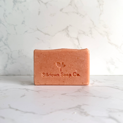 A rectangular bar of Silktown Soap Co. soap in a warm peachy-pink color, standing upright on a white marble surface with a matching marble background.