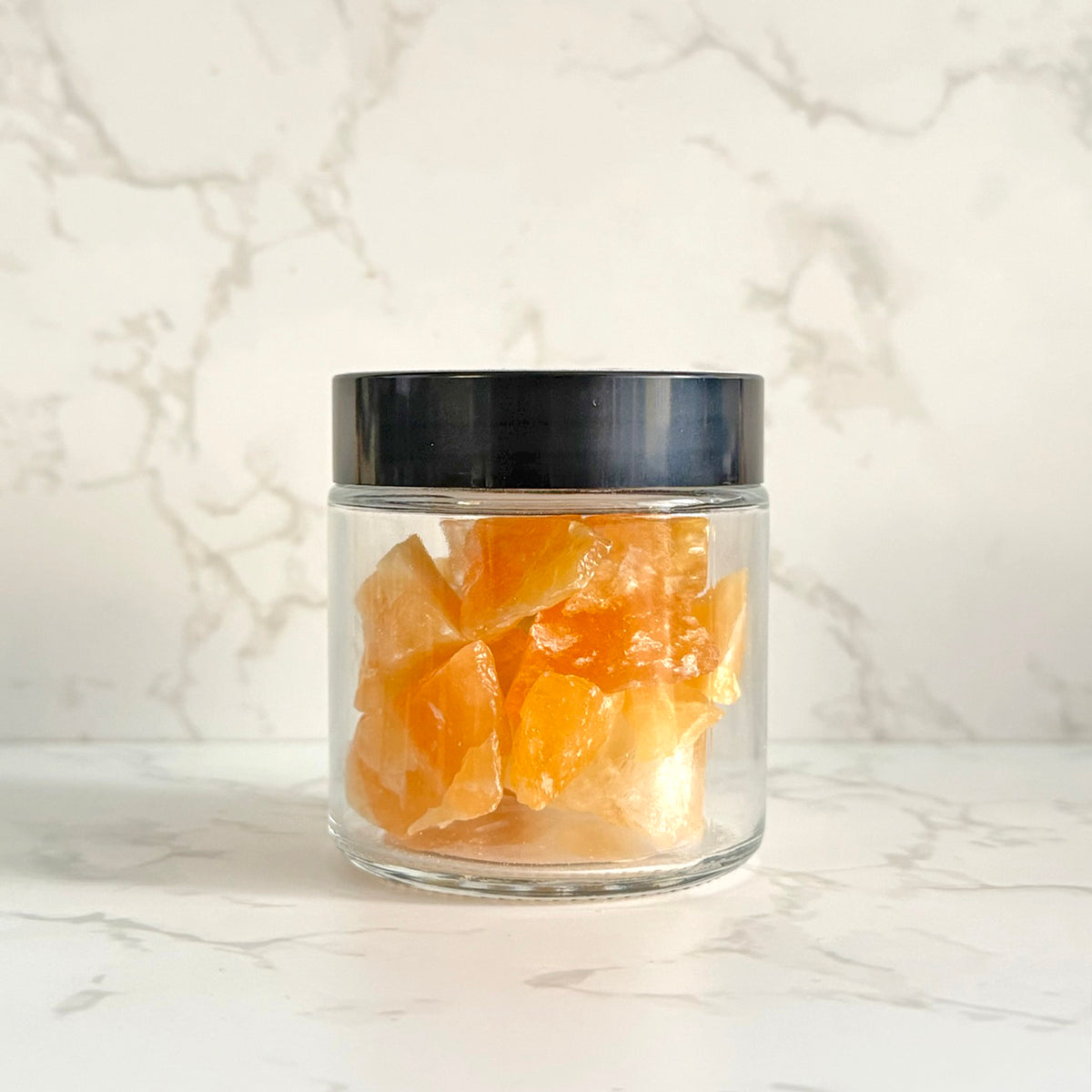 A clear glass jar with a black lid containing orange crystals, set on a white marble surface against a matching marble background.