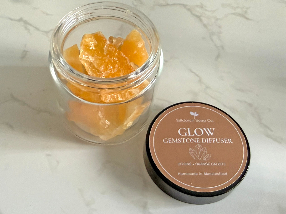An open clear glass jar filled with orange-yellow glow gemstone diffuser crystals sits beside a round brown lid labeled &