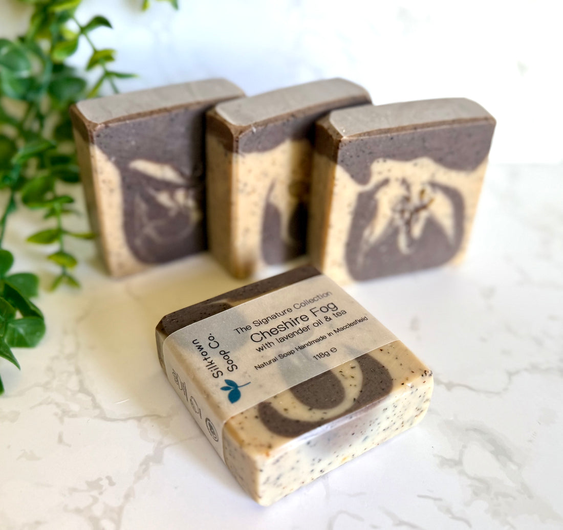 Three bars of soap with a packaging box on a light surface with greenery in the background