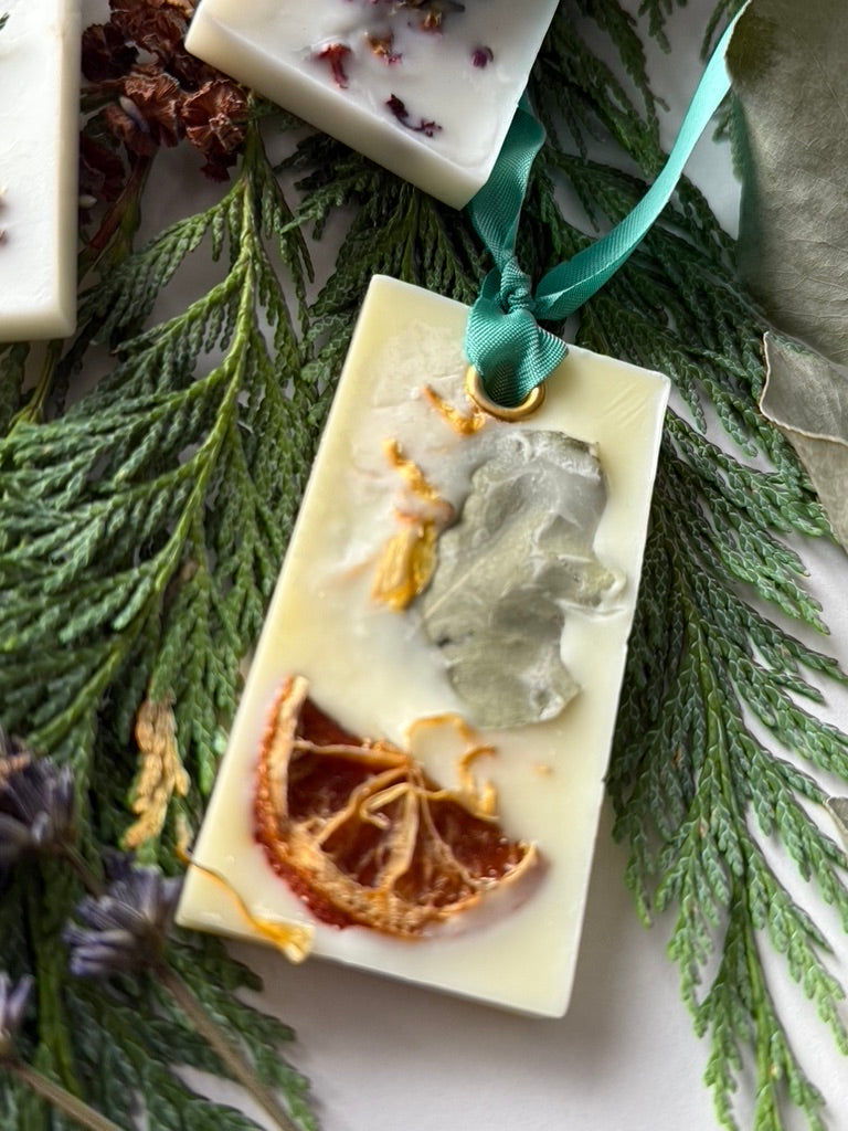 A natural wax freshener tablet with sections of dried citrus peel and botanicals, adorned with a green satin ribbon, placed on a bed of green faux foliage.
