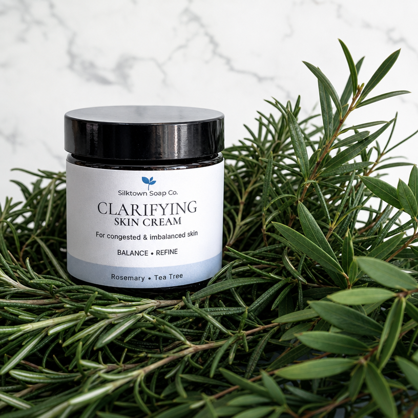 A jar of Silktown Soap Co. Clarifying Skin Cream sits nestled in fresh rosemary and olive branches against a white marble background.