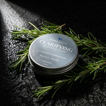 A round metal tin of Silktown Soap Co Clarifying Shaving Soap, surrounded by fresh green rosemary sprigs on a dark, textured surface with dramatic side lighting.