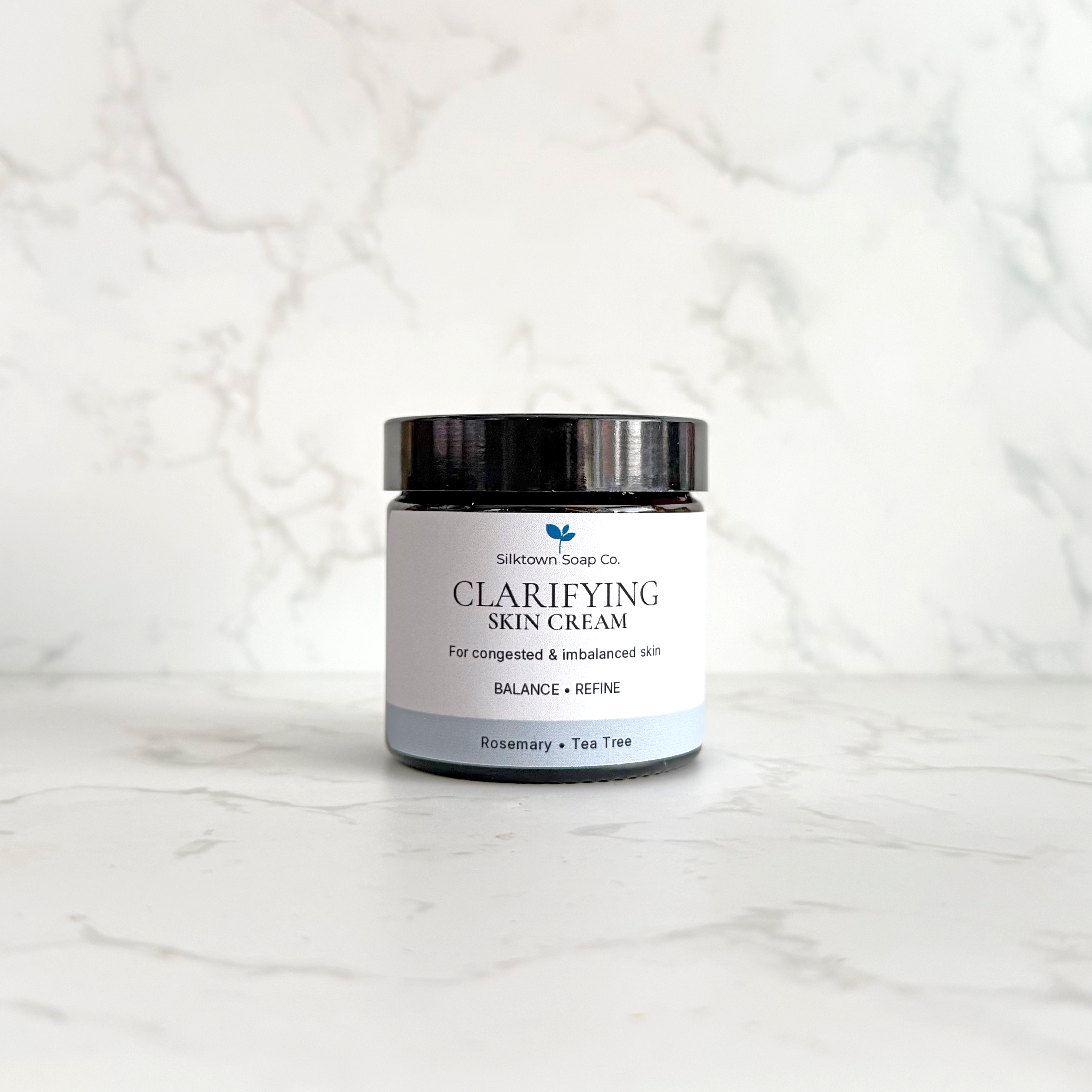 A jar of Silktown Soap Co. Clarifying Skin Cream with a black lid and white label, set on a light marble surface against a matching marble background.