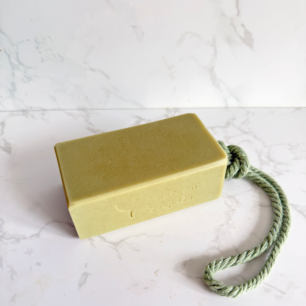 A rectangular bar of soap in a muted green color with a matching green rope loop, resting on a white marble-patterned surface.