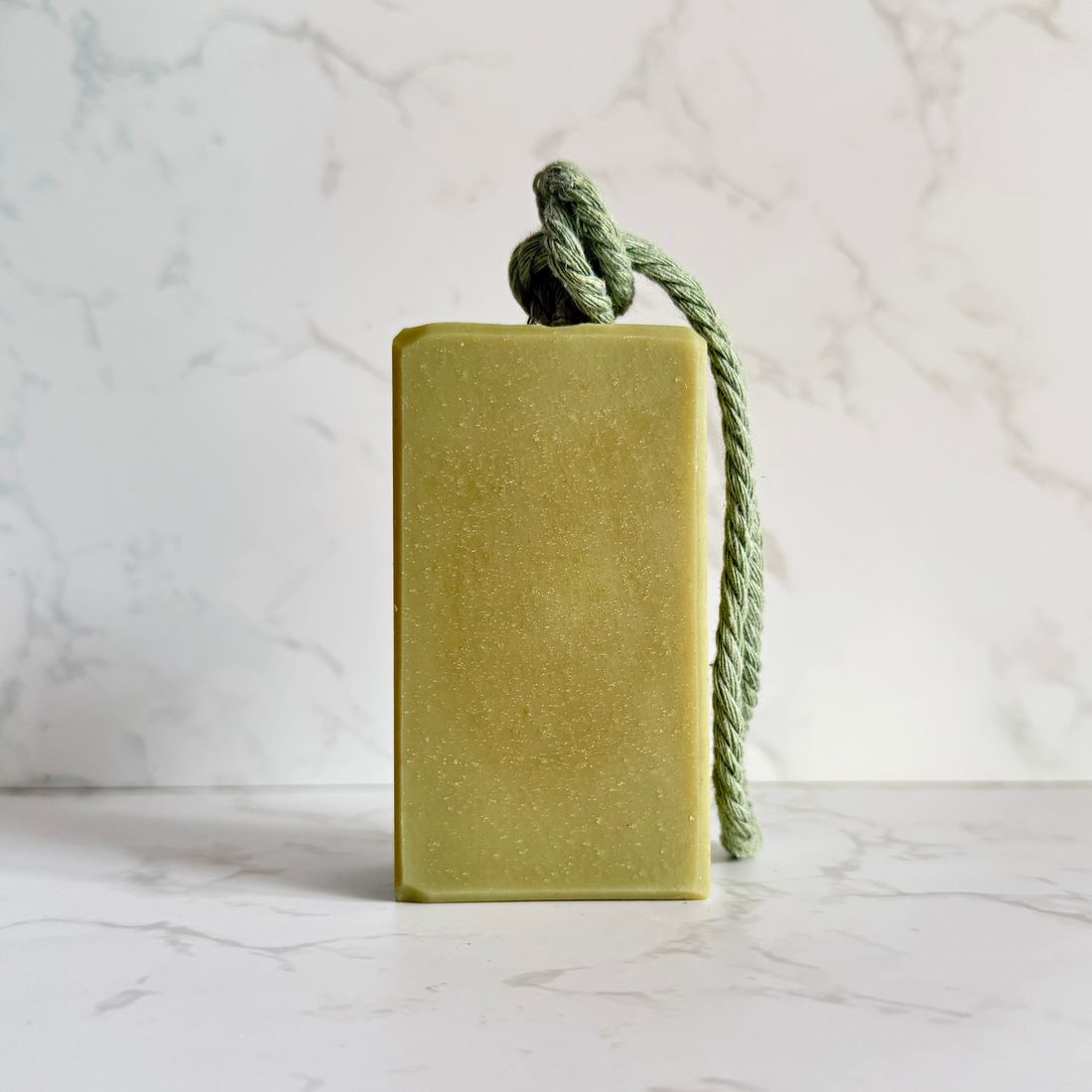 A rectangular bar of green soap  featuring a knotted sage green rope loop at the top, set against a white marble background.