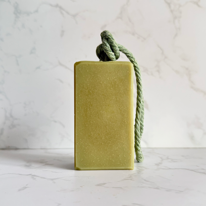 A rectangular bar of green soap  featuring a knotted sage green rope loop at the top, set against a white marble background.