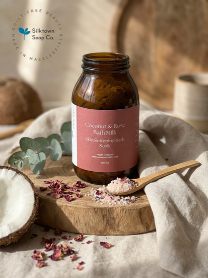 Bottle of Coconut &amp; Rose Bath Milk with a spoonful of bath salt on a wooden board, surrounded by coconuts and eucalyptus leaves.