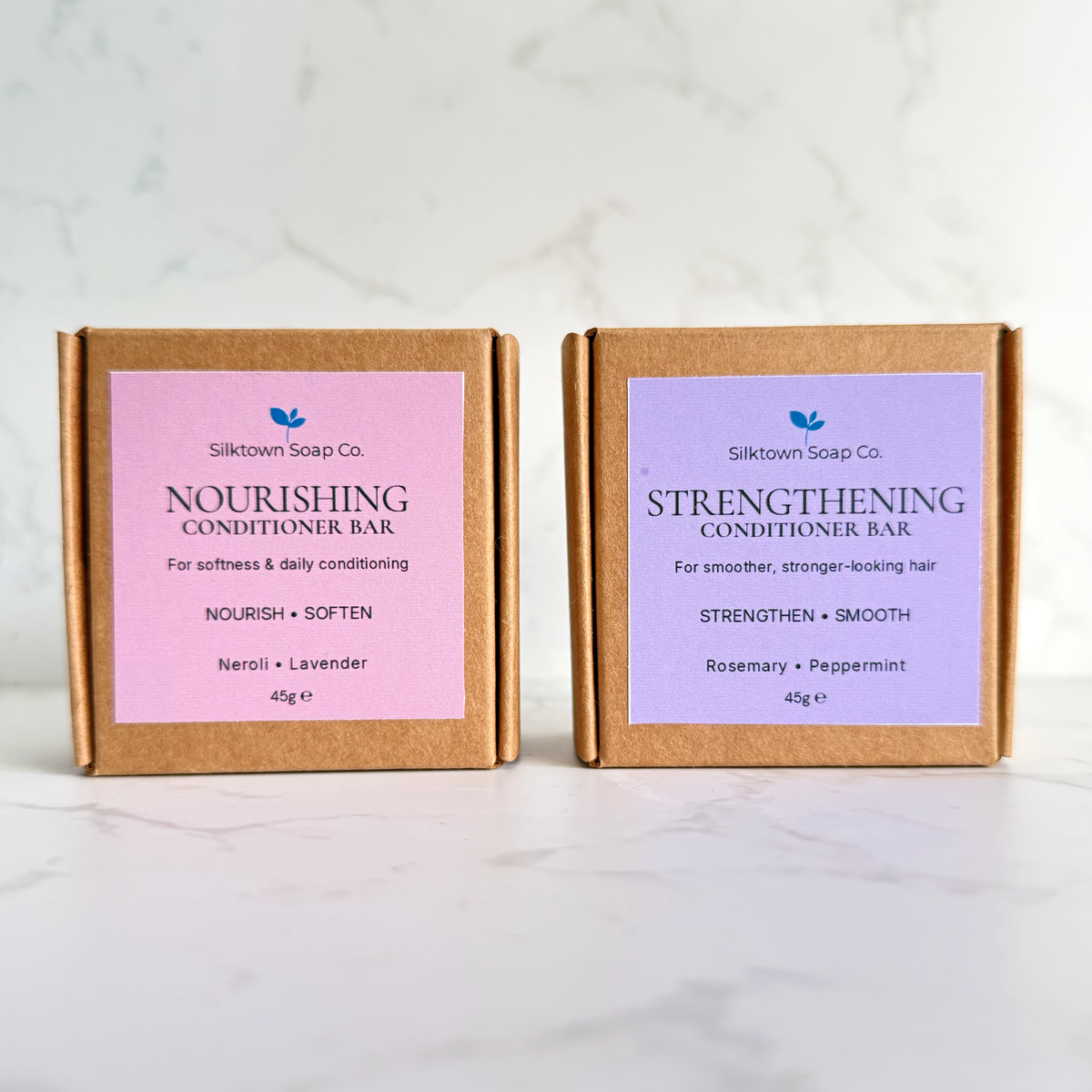 Two small kraft paper conditioner bars from Silktown Soap Co. stand side by side on a light marble surface against a white marble background.