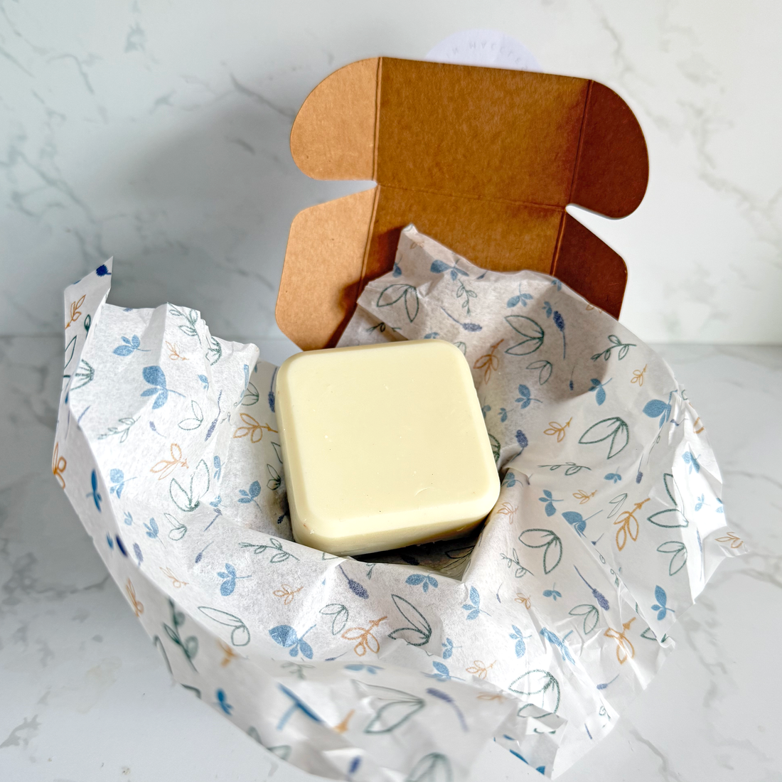 A square cream-colored conditioner bar nestled in white tissue paper with a blue and orange leaf pattern, sitting inside an open brown cardboard box on a light marble surface.