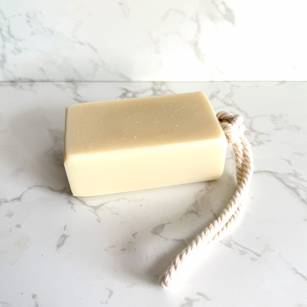 A rectangular bar of pale cream-colored soap with a natural beige rope loop, resting on a white marble surface with a matching marble background.