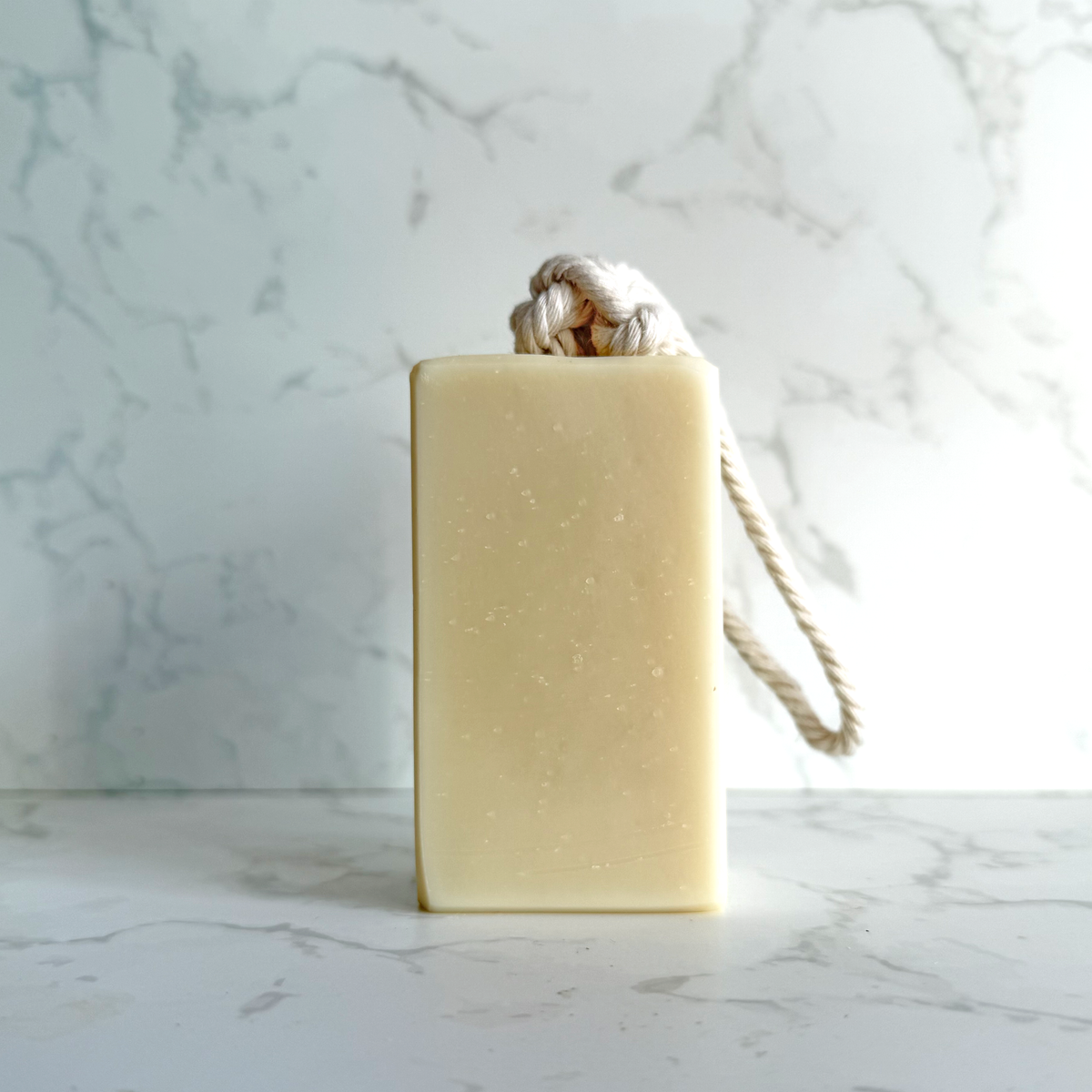 A rectangular pale cream soap bar with a knotted natural rope loop on top, standing on a light marble surface against a matching marble background.