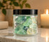 A clear glass jar with a black lid filled with green and pale yellow raw crystals, set on a marble surface with a blurred candle and plant in the background.