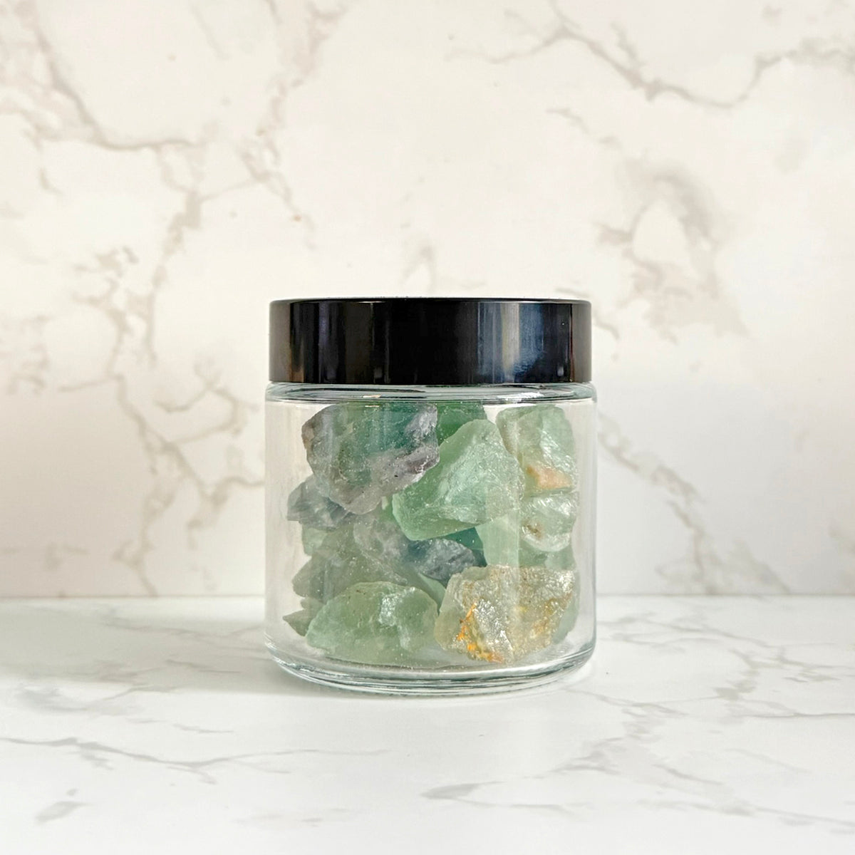 A clear glass jar with a black lid containing green translucent crystals, set on a white marble surface against a white marble background.
