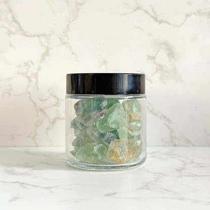 A clear glass jar with a black lid containing green translucent crystals, set on a white marble surface against a white marble background.