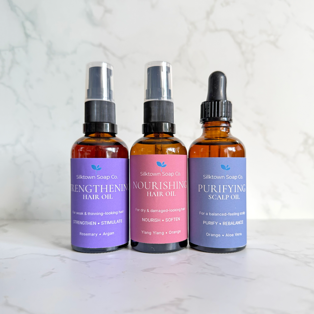 Three amber glass hair oil bottles from Silktown Soap Co. lined up on a white marble surface against a white marble background.