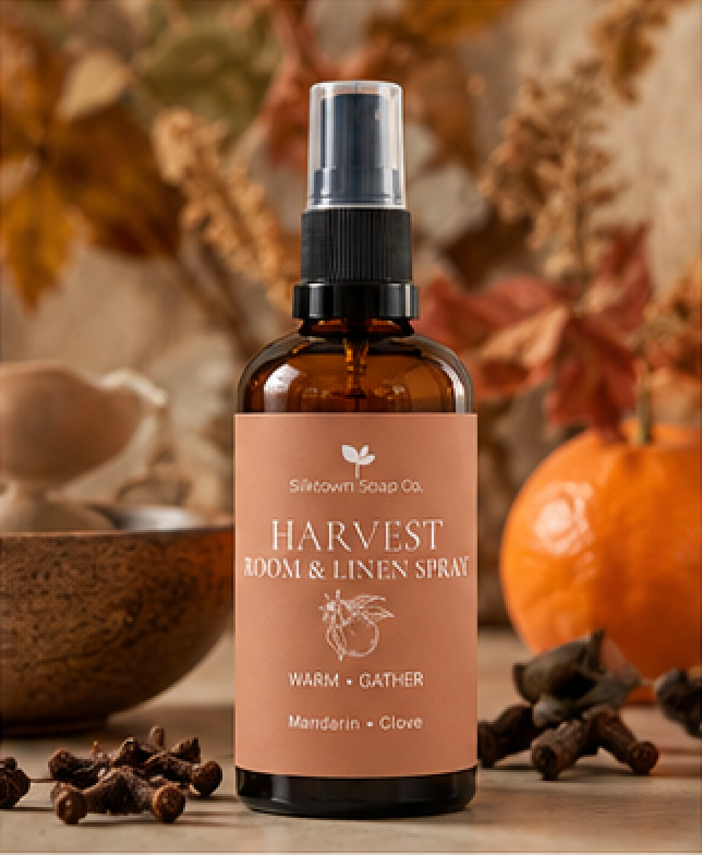 Silkworm Soap Co. Harvest Room &amp; Linen Spray in a warm amber glass bottle with a black spray top and a peach-colored label, styled on a rustic wooden surface with cloves, a bowl, and autumn leaves.