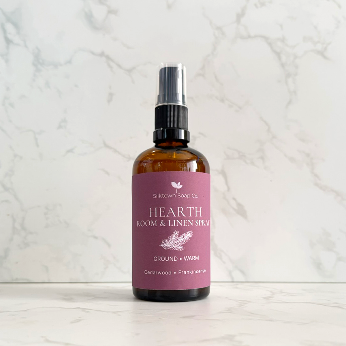 Silktown Soap Co. Hearth Room &amp; Linen Spray in an amber glass bottle with a dusty pink label, set against a white marble background.