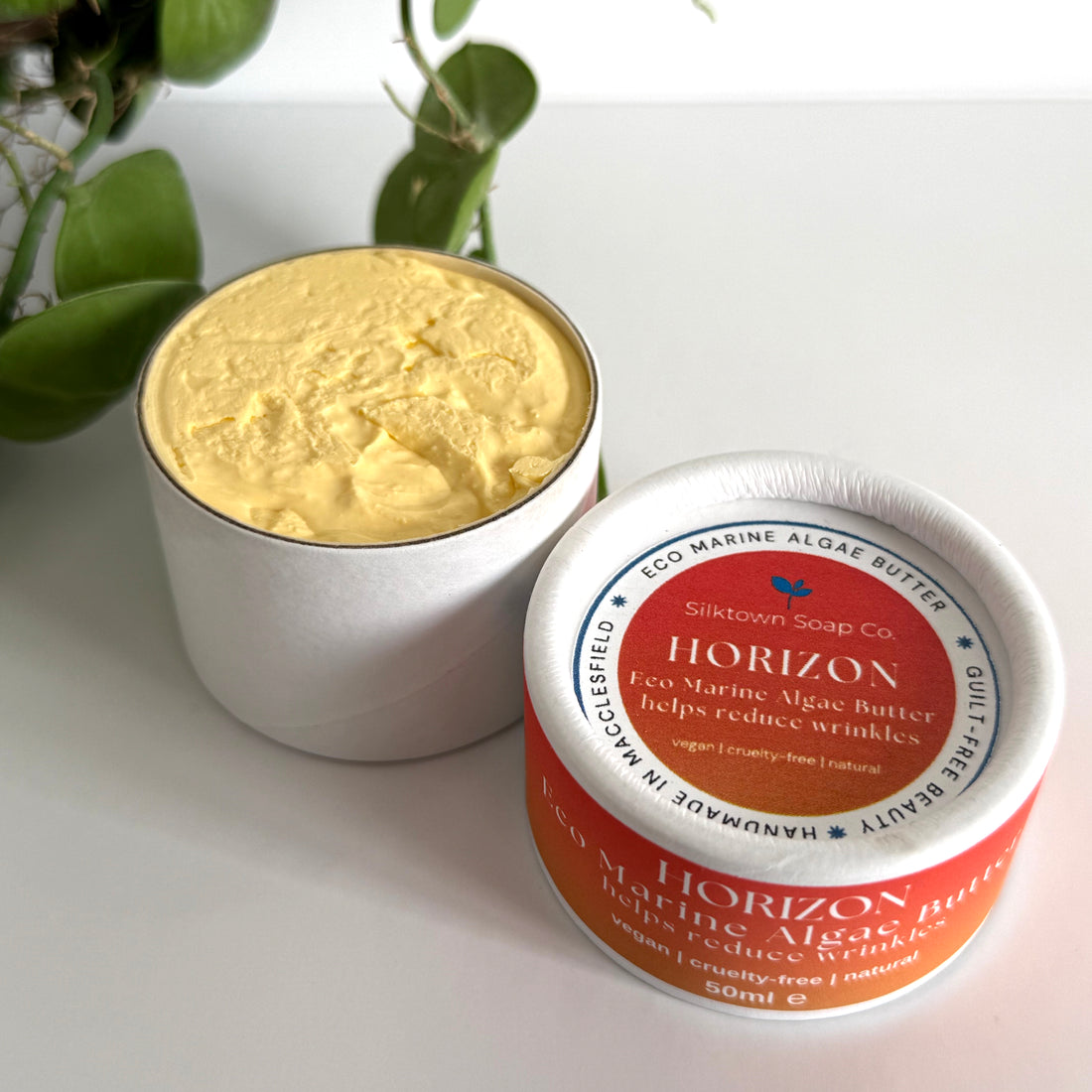 Jar of yellow cream with a red and white label on a light surface with green leaves in the background