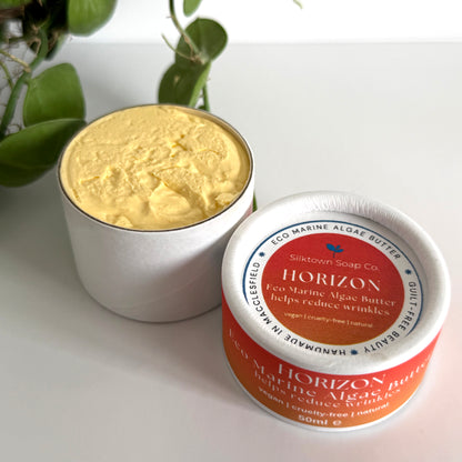 Jar of yellow cream with a red and white label on a light surface with green leaves in the background