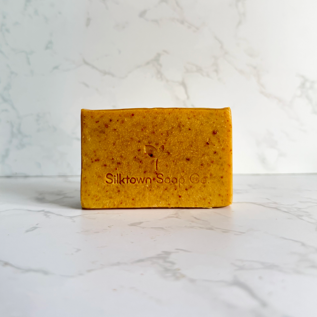 A rectangular bar of yellow-orange speckled soap standing upright on a white marble surface against a white marble background. The soap is embossed with the brand name &