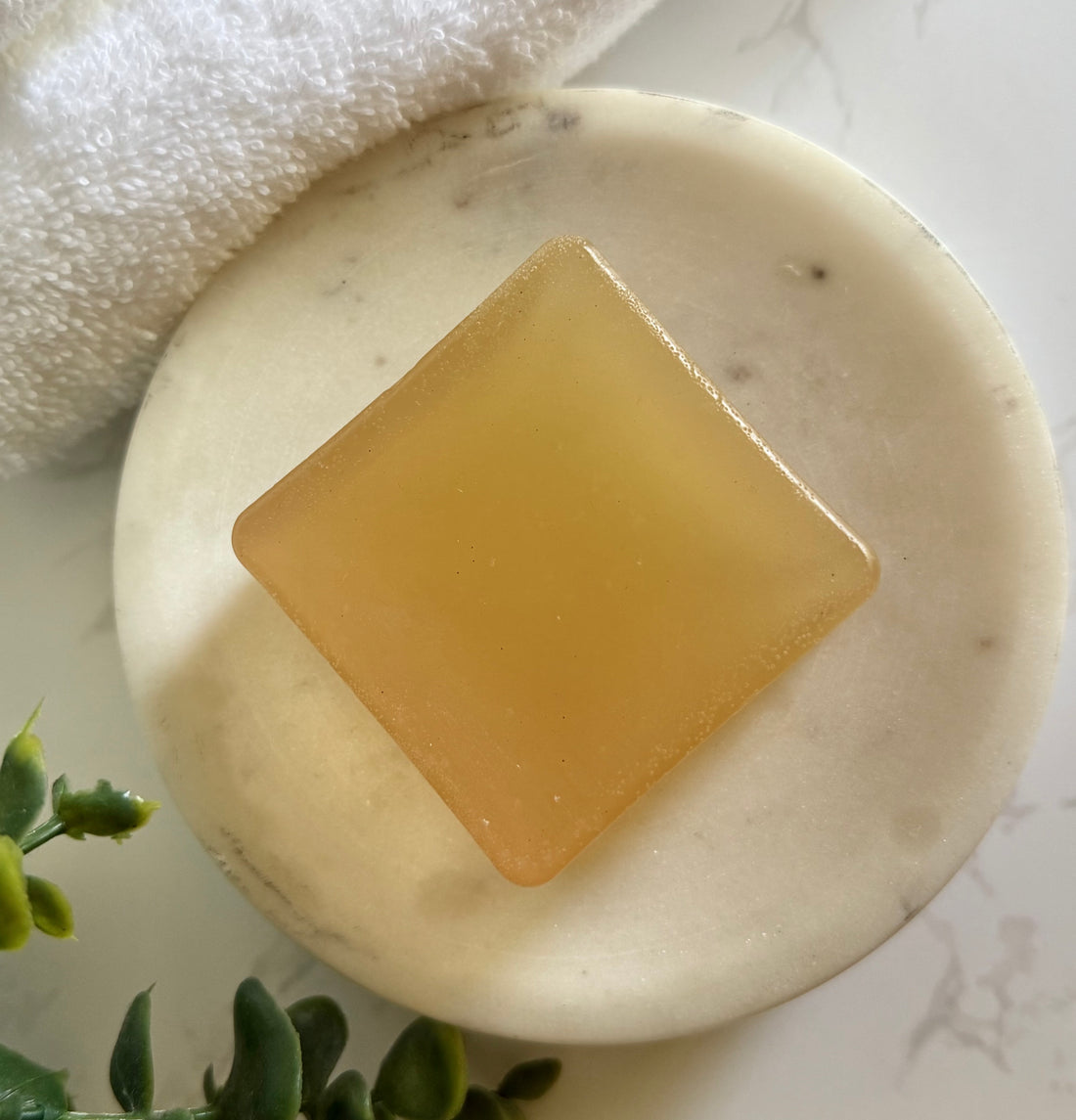 Bar of Luminary Natural Soap on a white marble surface with a towel and greenery in the background