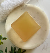 Bar of soap on a white marble surface with a towel and greenery in the background