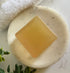 Bar of soap on a white marble surface with a towel and greenery in the background