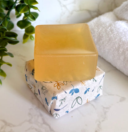Bar of soap on a box with floral wrapping, placed on a marble surface with a plant and towel in the background.