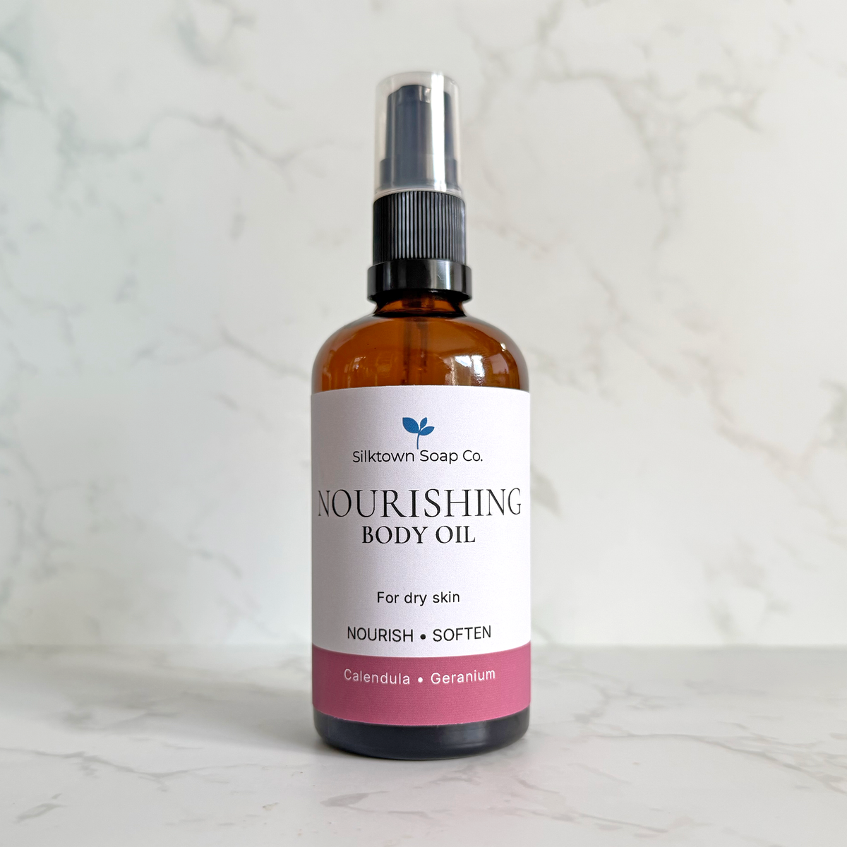 An amber glass spray bottle of Sliktown Soap Co. Nourishing Body Oil with a white label and pink bottom section, set against a light marble background.